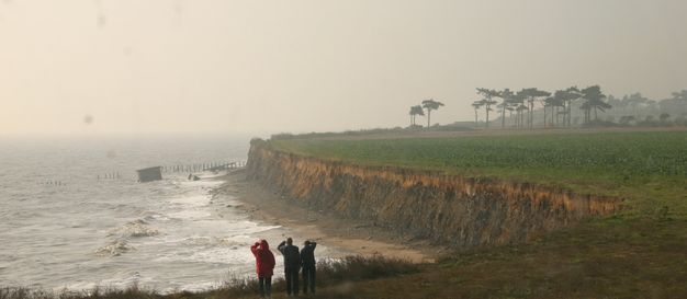 Three people standing in front of us look out at an expanse of steep cliffs that recede away from us. Waves lap at the foot of the cliffs.