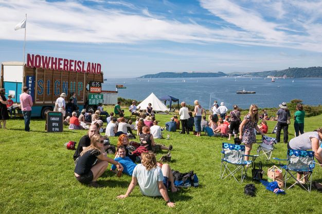 People gather on a grassy seaside near a stand with signs reading, “Nowhereisland”, “Sign up”, and “Embassy”.