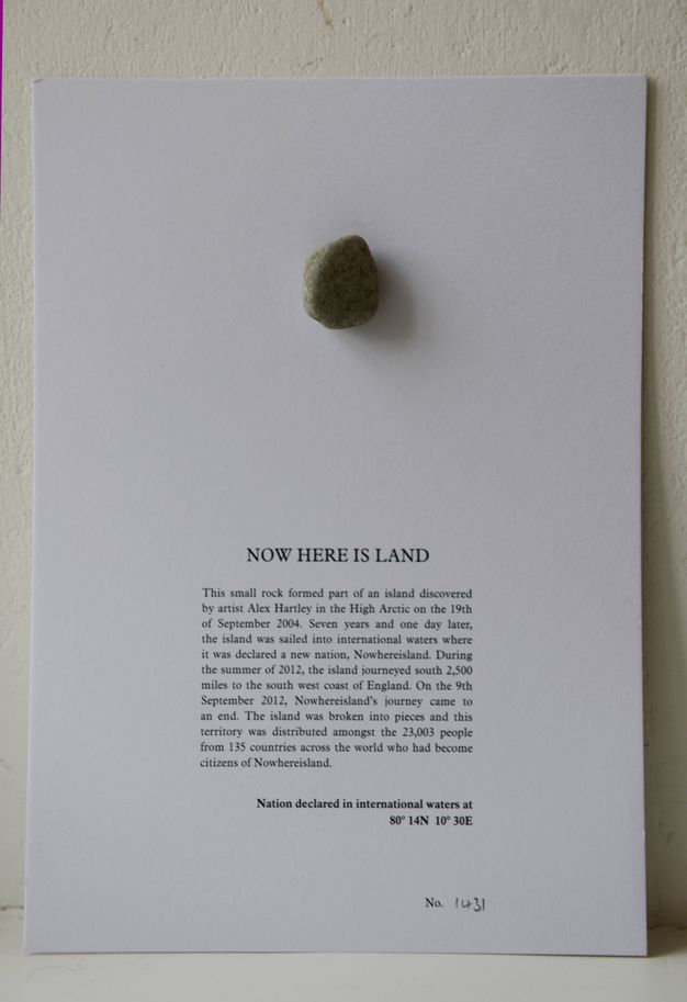 A dark pebble is affixed to a white card with text that begins, “Now Here Is Land”. Text continues below, and the card is numbered in pencil in the bottom right.