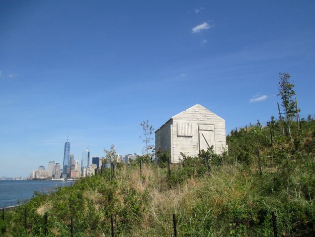 Installation. A plaster sculpture stands on a grassy hill with a view of the New York City skyline in the deep distance. The sculpture was cast using a wooden shed as a mould, so it captures the negative space inside the structure.