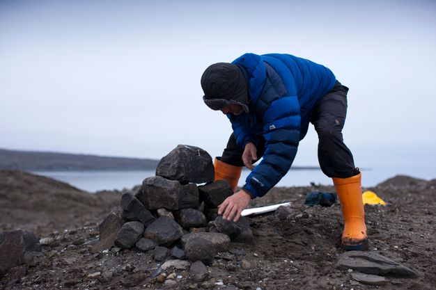 A man wearing a puffy blue coat and orange waders stands on a dark spit of land. He bends forwards and stacks rocks into a dome shape.