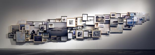 Installation. Framed photographs, maps, sketches, and other works on paper are hung in a tight grouping, many overlapping, to create a long, narrow shape that rises up the wall on which they are displayed.