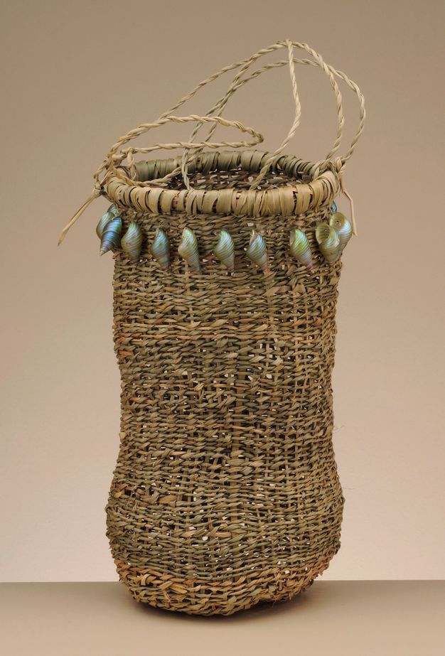 Basket. Tall woven vessel has a rounded bottom and iridescent, spiralling shells around the mouth. Thin handles loop up from the top.