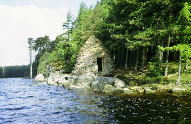 Architecture. A beehive-shaped stone hut has a rectangular opening. It is tucked into trees that come up to the water’s edge, where the hut sits.