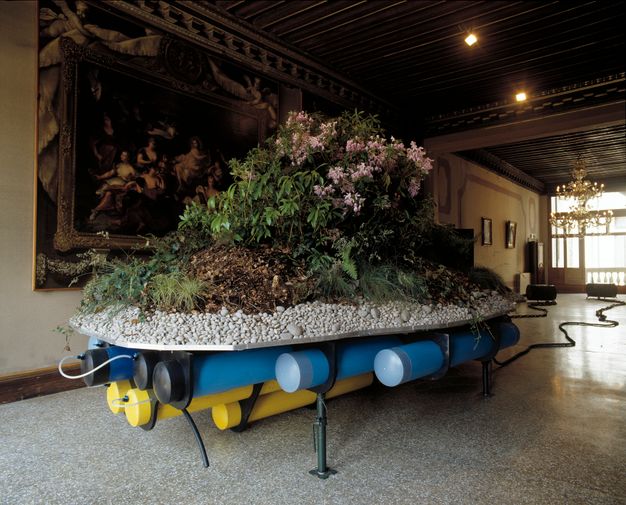Installation. A bank of rhododendrons sits on a mound of dirt supported by a layer of white rock. Blue and yellow pipes run under the tray holding the plants. The piece is set up in a gallery with wood ceilings, a brass chandelier, and stone floors.