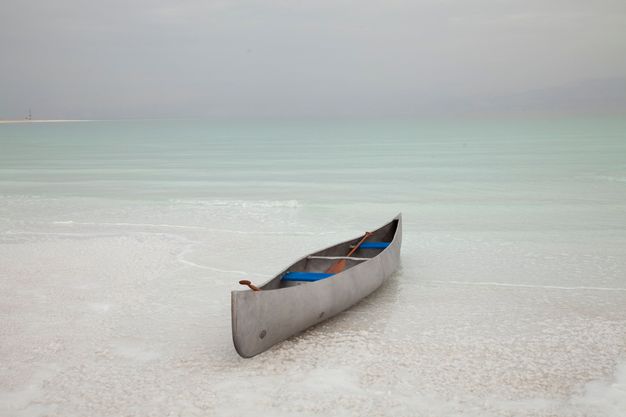 A silver-coloured canoe sits in the shallows of water that deepens from ivory white close to us to pale glacial green in the distance. The horizon is hazy lavender blue.