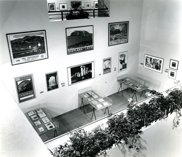 Installation. Black and white photograph overlooking a double-high room hung with eleven posters and works on paper. Three cases along the far wall are filled with open books. A planter in front of us is filled with greenery, which separates us from the space below.