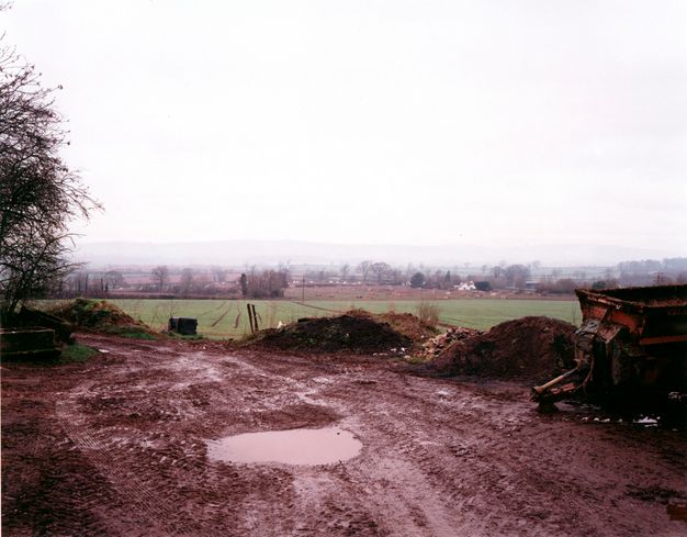 A construction vehicle is parked along the edge of a muddy area pocked with puddles. A green field beyond leads back to a town on a hazy, overcast horizon.