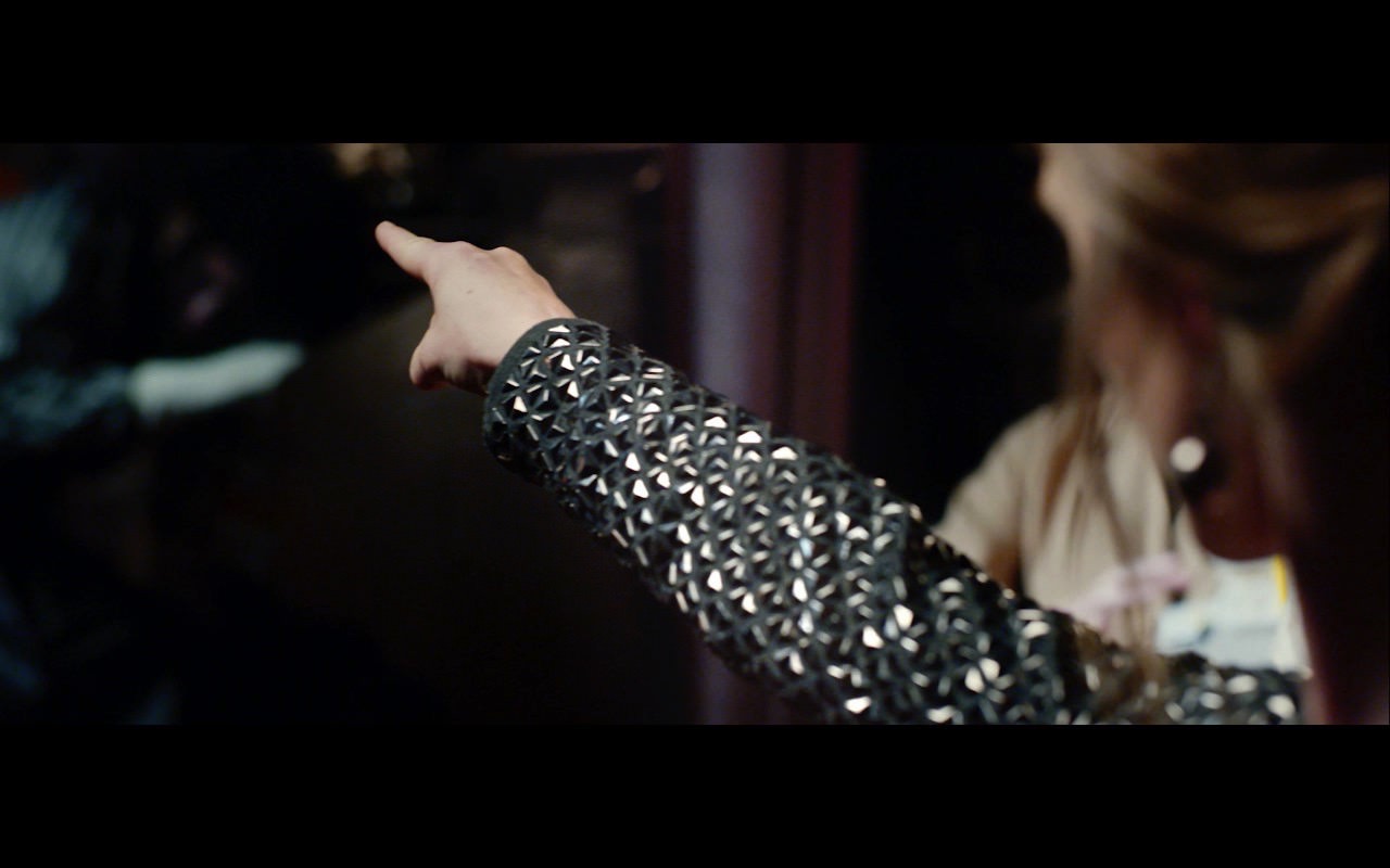 We look along the shimmering black and silver-clad arm of a woman who points towards a shadowy background.
