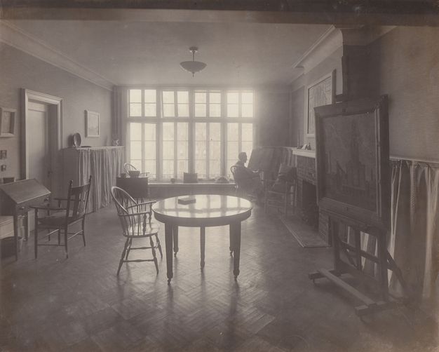 Black and white photograph. A round table and one chair is backlit by a wall of windows at the far end of this room. A desk, easel, and other furniture lines the walls. A sitting person is silhouetted near the windows.