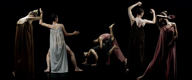 Six dancers wearing long, flowing gowns pose with arms and legs flexed or rigid against a black background.