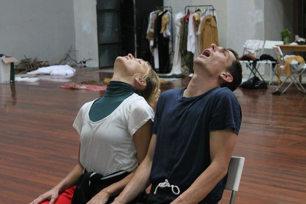 Colour photograph. A man and a woman sit side by side in a rehearsal space. Both throw their heads back so we look on the undersides of their chins. The man’s mouth is wide open.