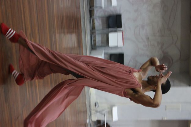 Colour photograph. A dancer steps forwards while leaning his torso back, creating a long diagonal. Both hands are raised with forefingers and thumbs pinched in front of his eyes. A swath of the rose-pink garment he wears flutters behind him.