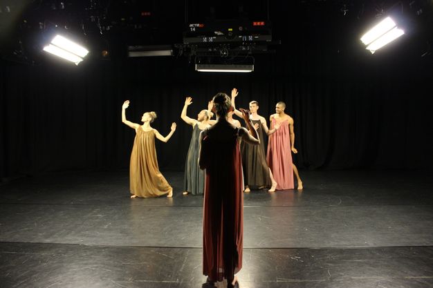 Colour photograph. A woman close to us stands facing away, addressing a row of five dancers. All six people wear long, flowing garments in different colours. Bright light from the sides, within frame, makes the dancers stand out in stark contrast against the black background.
