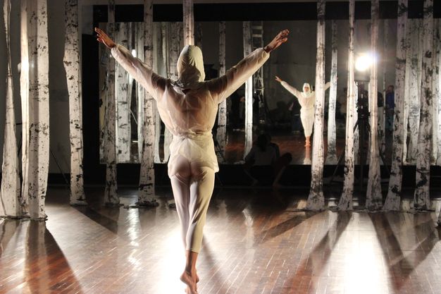 A dancer balances on his tip toes, both arms stretched out and up. He wears a white hood and clothing. He faces away from us and is reflected, along with the filming crew, in a mirror on the wall opposite us. Strips of white fabric hanging from the ceiling are streaked with dark grey.
