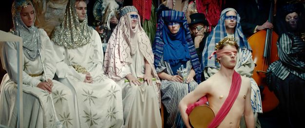 Seven people wearing patterned robes sit in a row, with more behind. In front of them, a young bare-chested man holds a gold drum. He wears a pink blindfold and sash, and a gold crown of leaves and gold discs on his nipples.