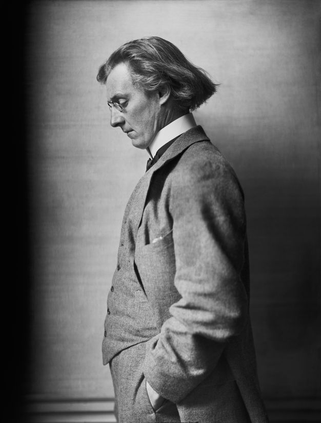 Black and white photograph. Shown from the hips up, a cleanshaven man with chin-length hair and pale skin stands facing our left in profile. He looks down through pince-nez, and the hand we can see is in his pocket. His three-piece suit has a high white collar.