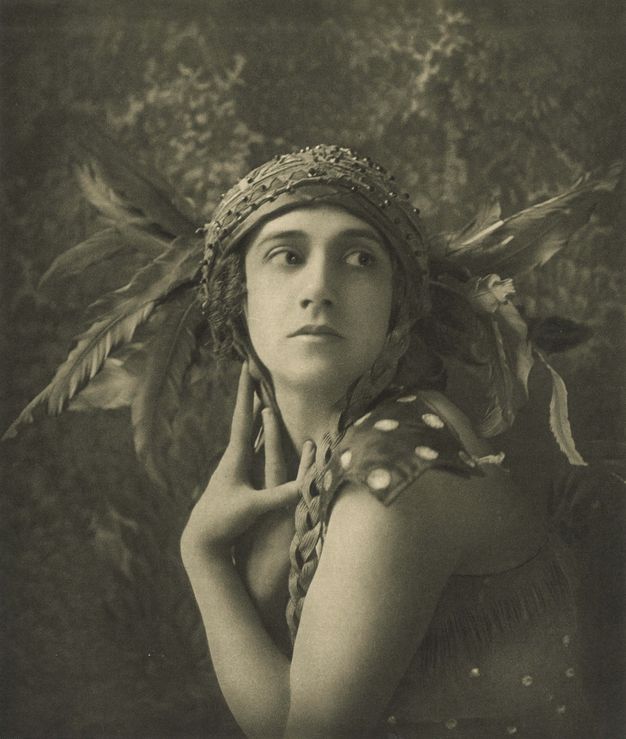Black and white photograph. Shown from the waist up, a woman wearing a feathered headdress poses with her body facing our left as she turns her head to look off to our right. Both hands lie along her heck. The feathers fan out from the sides of her head like wings. Beaded silk wraps across her head like a turban.