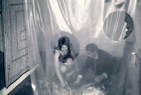 Black and white photograph. Holes are cut out of a sheet of translucent plastic hanging between us and visitors beyond. A man and a woman crouch and both reach for something on the floor.