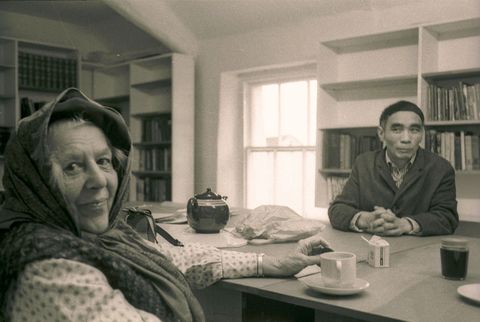 Black and white photograph. Two people sit at a table in a shelf-lined room. Closer to us, a pale-skinned woman’s head is wrapped in a scarf. She looks at us and smiles, one hand resting on the table. Across the table, a medium-skinned man sits with his fingers interlaced and slightly smiles, lips closed, at the woman.