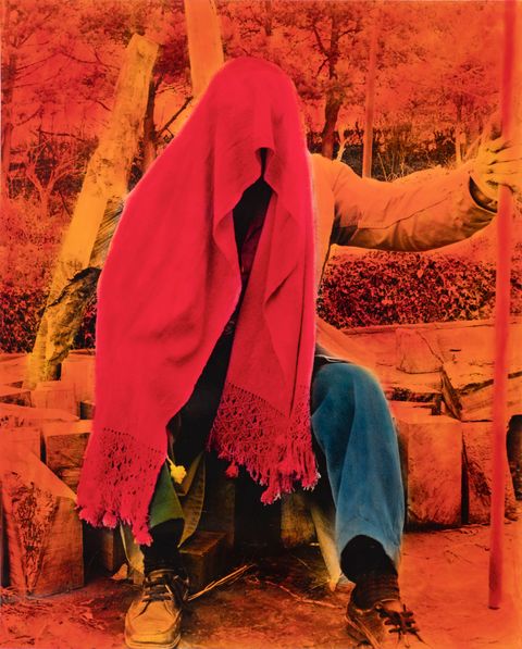 Hand-coloured black and white photograph. A person sits with a scarlet-red scarf draped over their head, obscuring their face. One hand is out and holds a staff. The background is painted traffic-cone orange and the pants indigo blue.