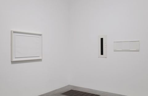 Installation. Three works of art are hung in the corner of a gallery. A rectangular piece to our left and a long, narrow piece to our right are totally white. The third has a black stripe down a white support or background.