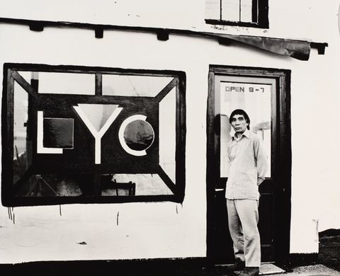 Black and white photograph. A man stands in front of a door, hands behind his back, looking at us. Text applied to the glass in the top half of the door reads “Open 9-7”. The storefront window to our left has the initials “LYC” in a geometric design. The L nestles a square in its bend, the Y a triangle, and the C a circle.