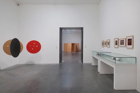 Installation. To our right, six framed works of art hang over two long cases with accordion-folded strips of paper in this gallery. Three discs, one red, one black, and one fawn brown, are suspended from the ceiling to our left.