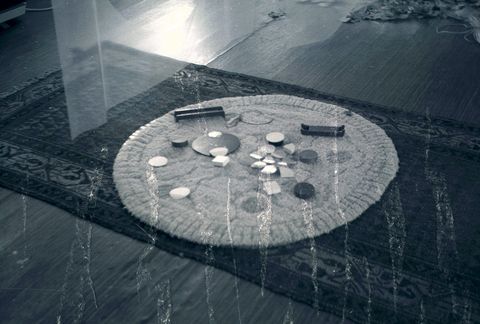 Black and white photograph. Discs are laid out on a round area rug placed on top of a rectangular area rug. Texture on the surface of the photograph flickers like flames across the bottom of the image.