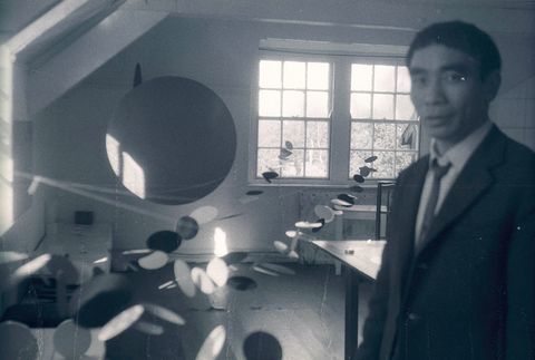Black and white photograph. A person, Li Yuan-chia, stands close to us in his studio. He is blurry because the camera’s focus is on the end of a long path of fluttering discs that lead back to windows on the far side of the room.