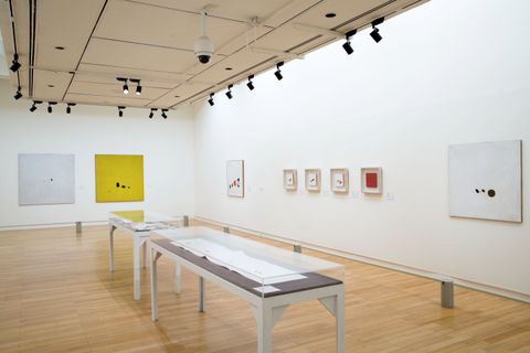 Installation. Two long cases with works on paper are in the middle of a gallery hung with works of art. Most of the compositions are square and dominated by white, yellow, or red, which act as backgrounds for small spots and marks in black or other colours.
