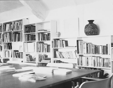Black and white photograph. Book-lined shelves fill the wall beyond a conference-sized study desk.