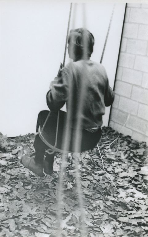 Black and white photograph. A person swings away from us near a corner. The ground is covered in leaves.