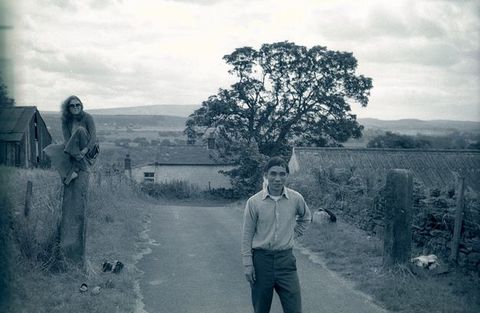 Black and white photograph. A man stands in a road and a woman sits on a high post a short distance from us. Both look at us and smile. The road slopes down as it moves away, so we look onto the roofs of structures in front of a flat expanse.