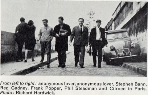 Black and white photograph. Four men walk towards us on a street past a parked car. A man and a woman with their arms around each other walk away from us, to our left.