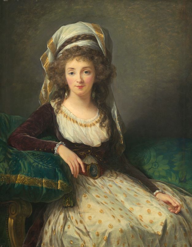 Painting. Shown from the knees up, a pale-skinned young woman wears a short wine-red jacket over an ivory-white gown. She looks out at us with blue eyes under faint brows. A gold-edged white turban nestles on a cloud of curls, and she wears a cameo on her belt.