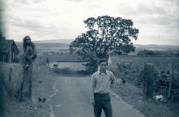 Black and white photograph. A man stands in a road and a woman sits on a high post a short distance from us. Both look at us and smile. The road slopes down as it moves away, so we look onto the roofs of structures in front of a flat expanse.