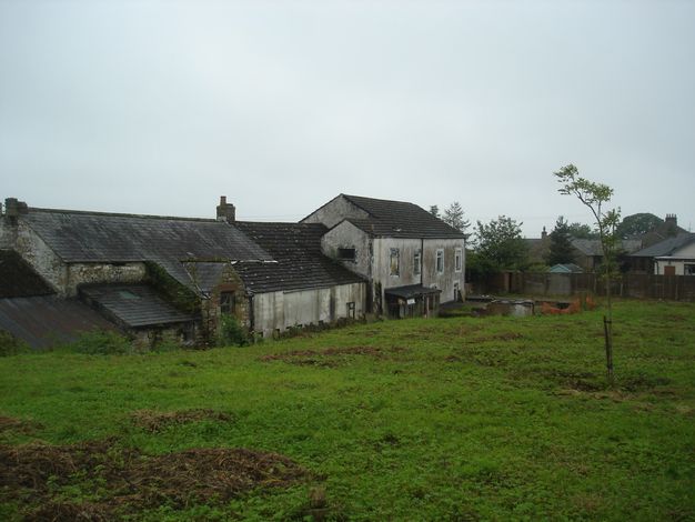 Architecture. Grey-stained white structures are across a weedy lawn. A long, warehouse-like building is attached to what could be a two-storey house.