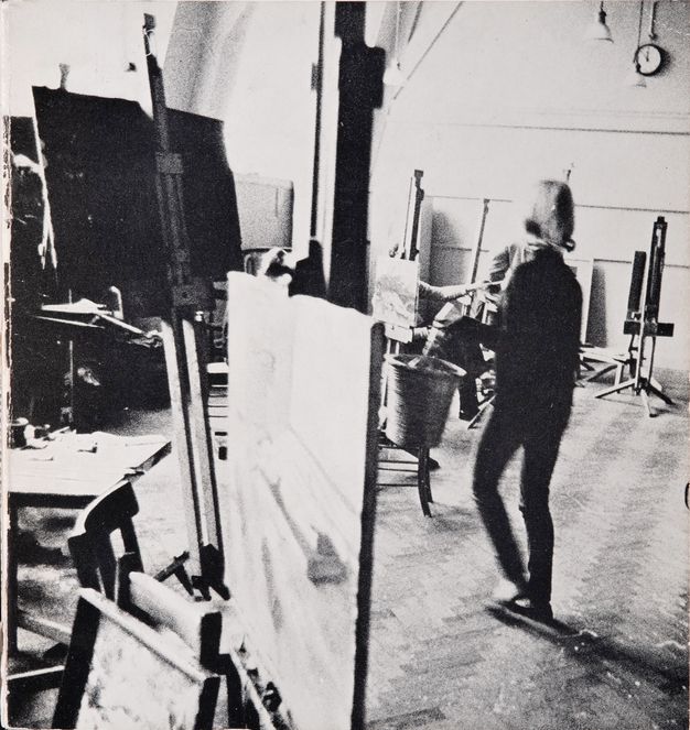 Black and white photograph. Two people in a studio space with paintings on easels. One person is blurry as they walk across the space.