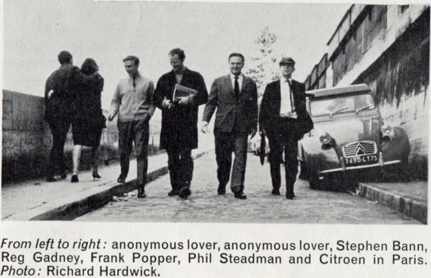 Black and white photograph. Four men walk towards us on a street past a parked car. A man and a woman with their arms around each other walk away from us, to our left.