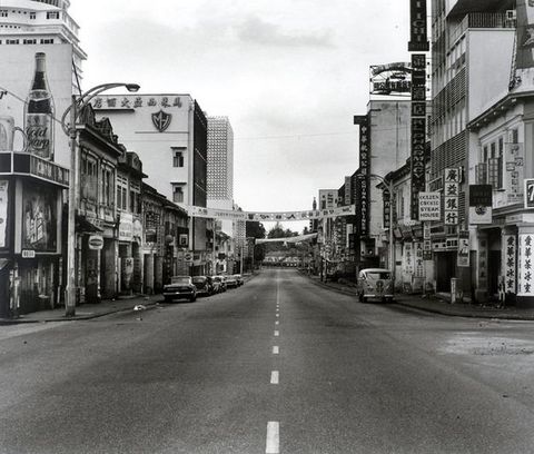 Black and white photograph. An empty two-lane road is lined with buildings hung with signs in Chinese and English. Cars line the curb to our left. A single Volkswagen bus is parked facing us to our right. Two banners are hung from buildings to span the street.