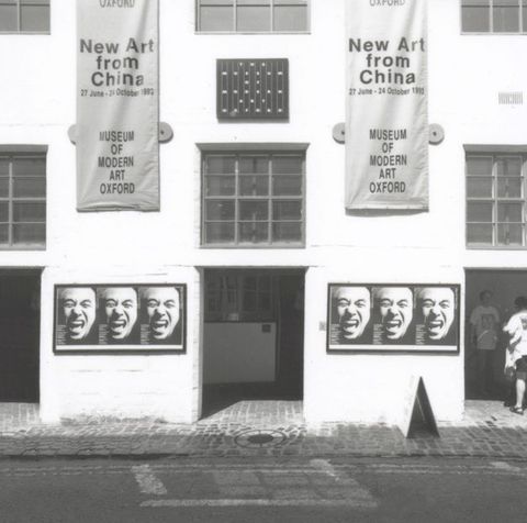 Black and white photograph. Two banners hanging on a white building have the title and location of the exhibition along with the dates, “27 June – 20 October 1993”. Below each hangs a panel showing a close up of a laughing face three times. People enter a doorway to our right.