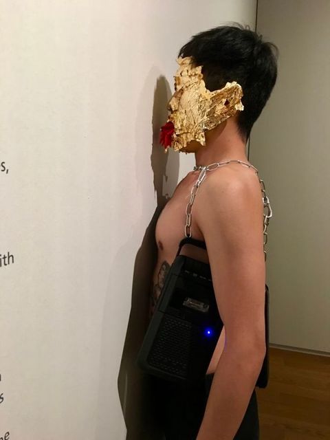 Performance still. A medium-skinned, bare-chested man presses his torso to a gallery wall. Cassette players hang from chains around his chest, and his face is covered with gold leaf. Red chili peppers flick out of his mouth like thin tongues.