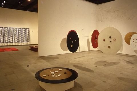 Installation. At least eight round discs, like tabletops, float suspended from a gallery ceiling. A seventh sits on a pedestal and holds round stones or objects made of another material. The hanging discs are painted brown, red, or black. A sculptural piece hanging on the wall seen through a doorway to our left is of ladder or trellis-like structures creating a dense geometric pattern.