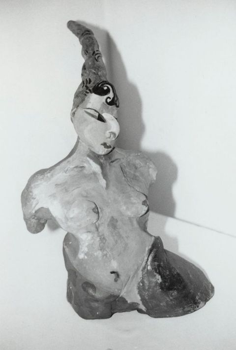 Sculpture. Black and white photograph of the torso and head of a stylized woman. A thin cone-like hat or headdress is over dark hair. Some of her features are exaggerated, like a curving, half-moon nose.
