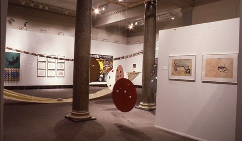 Installation. Three round disks hang from the ceiling between two polished marble columns in a gallery. Works of art and sculpture hang on the floating walls and occupy the space within.
