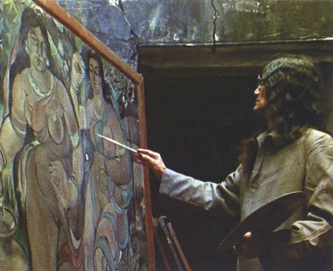 Video still. A medium-skinned man with long black hair painting a canvas showing two life-sized, stylized, medium-skinned women. He holds a round palette in the hand closer to us.