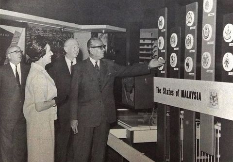 Black and white photograph. A man wearing glasses and smiling holds out a hand to gesture towards a display illustrating “The States of Malaysia”. Two other men and a woman look on, two of them also smiling. The men wear suits, and the woman a pillbox hat and gloves, with a skirt suit.