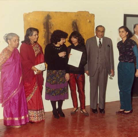Five women and a man with medium skin stand in a row in a gallery hung with works of art.