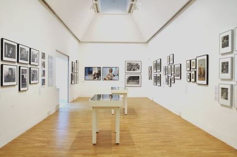 Installation. Framed black and white and colour photographs and perhaps other works on paper hang along the three walls of a gallery stretching before us. Two cases are in the middle of the space.